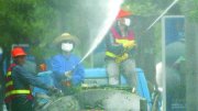 Workers spray water from a truck, wearing masks and hats, in a misty urban setting.