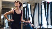 A dancer in a black leotard poses gracefully in a bright studio, with mirrors reflecting her movement.