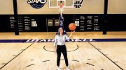 Katie Benzan stands on a basketball court holding a ball, with a hoop in the background.