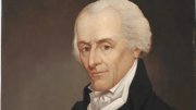 Portrait of a man with white hair, wearing a black coat, arms crossed, thoughtful expression.