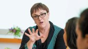 A woman in glasses gestures while speaking to two attentive listeners at a table.