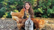 Alene Anello smiling surrounded by four chickens in a natural outdoor setting.
