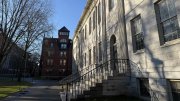 University Hall building with steps, shadows of trees, and a red brick structure in the background.