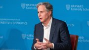 A man in a suit speaks from a chair, with a blue Harvard backdrop.