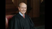 A smiling judge wearing a black robe, seated in a wood-paneled courtroom.