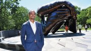 Brandon Terry, wearing a blue suit, standing before The Embrace, a large bronze sculpture of intertwined arms in Boston Common.