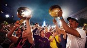 A crowd of people shout and march during a nighttime demonstration, while a man and woman in the foreground hold two silver-colored pans above their heads and bang on them with sticks