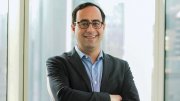 Matt Levine in a dark blazer and glasses stands smiling with arms crossed in front of a large window in a city building.