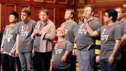 A diverse group of individuals standing on stage, wearing matching shirts and smiling.
