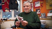 Older man in a green sweater holds a postcard in a warmly decorated office.