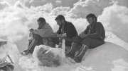 Three climbers seated on a snowy summit, surrounded by clouds, appearing contemplative.