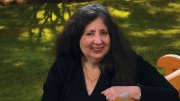 Jane Rosenzweig seated on a wooden bench in a grassy outdoor setting, smiling warmly.