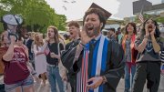A jubilant graduate shouts into a megaphone, surrounded by a cheering crowd.