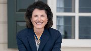 Anne Neal Petri in a navy suit leans on a wooden chair against an exterior wall of Mount Vernon..