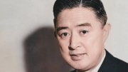 Man in a suit holding a pen, smiling, seated at a desk with a soft background.
