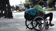 A man in a wheelchair with a rolled-up sleeping bag strapped to the back