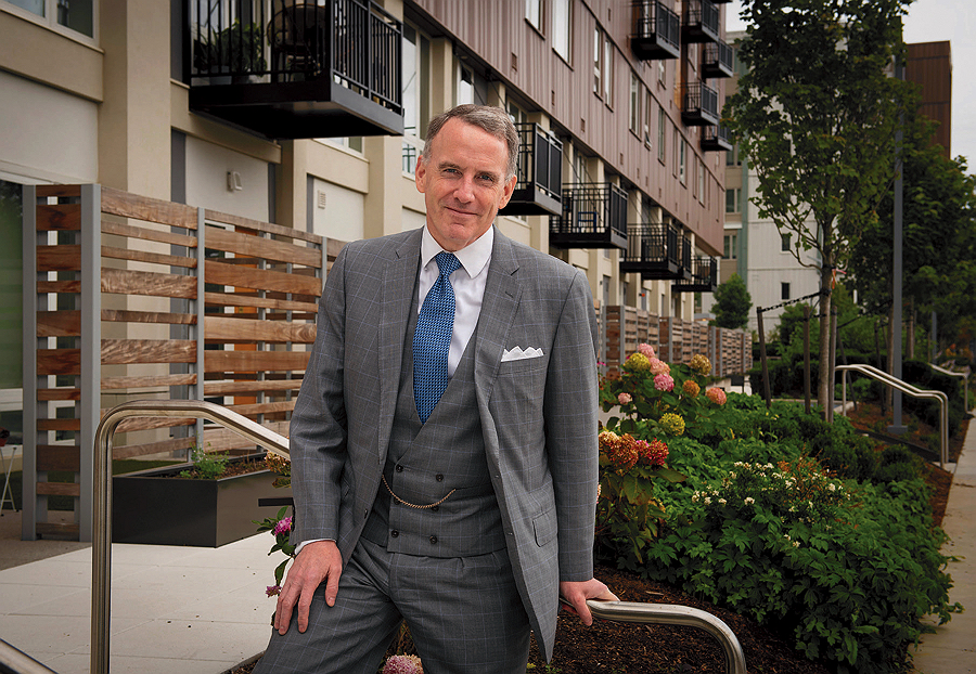 Man in a gray suit standing in front of a building and greenery.