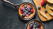 Two small cast iron pans with berry-topped desserts, dusted with powdered sugar, alongside lemon slices.