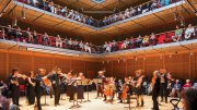 A lively concert in a modern auditorium with an audience seated on multiple levels.