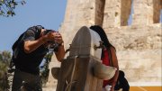 Man splashing water on his face at outdoor fountain beside woman holding cup near stone building.