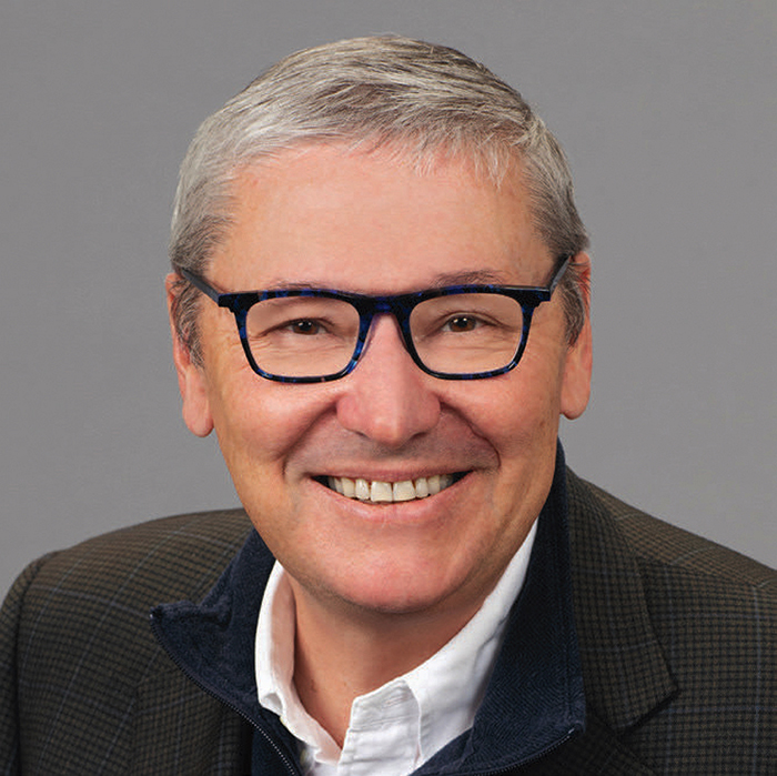 N. Gregory Mankiw, Smiling with short gray hair and glasses wearing a dark jacket over a white collared shirt.