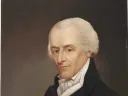 Portrait of a man with white hair, wearing a black coat, arms crossed, thoughtful expression.