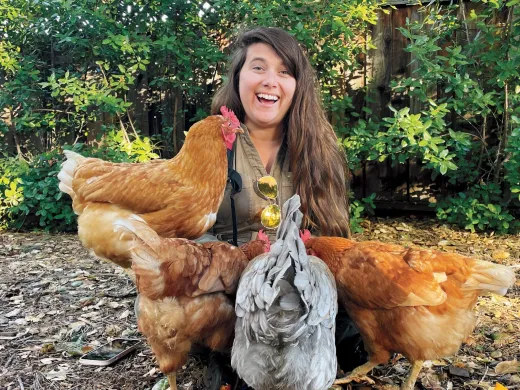 Alene Anello smiling surrounded by four chickens in a natural outdoor setting.