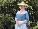 Woman in historical dress standing in front of green foliage, smiling brightly.