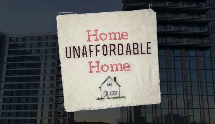 Cross-stitched fabric with the text "Home Unaffordable Home" over a background of high-rise apartment buildings.