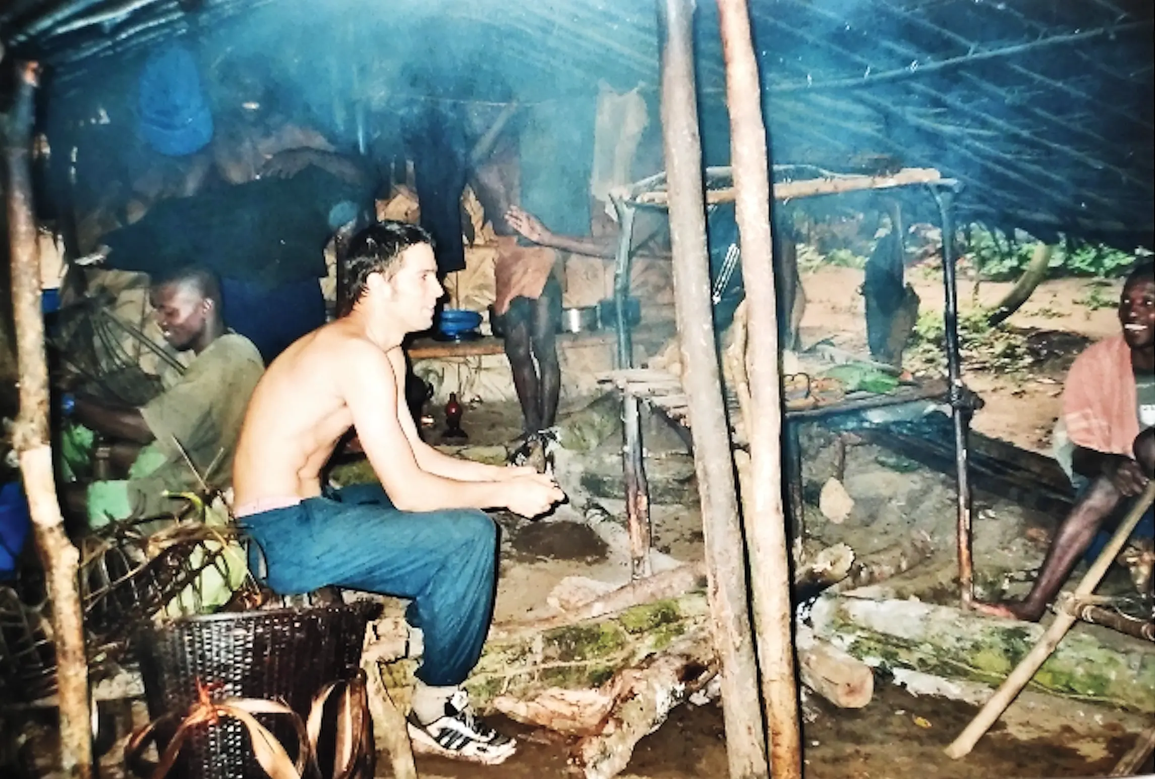 People sitting inside rustic forest shelter with firewood and cooking area.