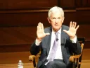 A man with gray hair gestures with both hands while seated on a wooden stage.