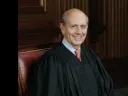 A smiling judge wearing a black robe, seated in a wood-paneled courtroom.