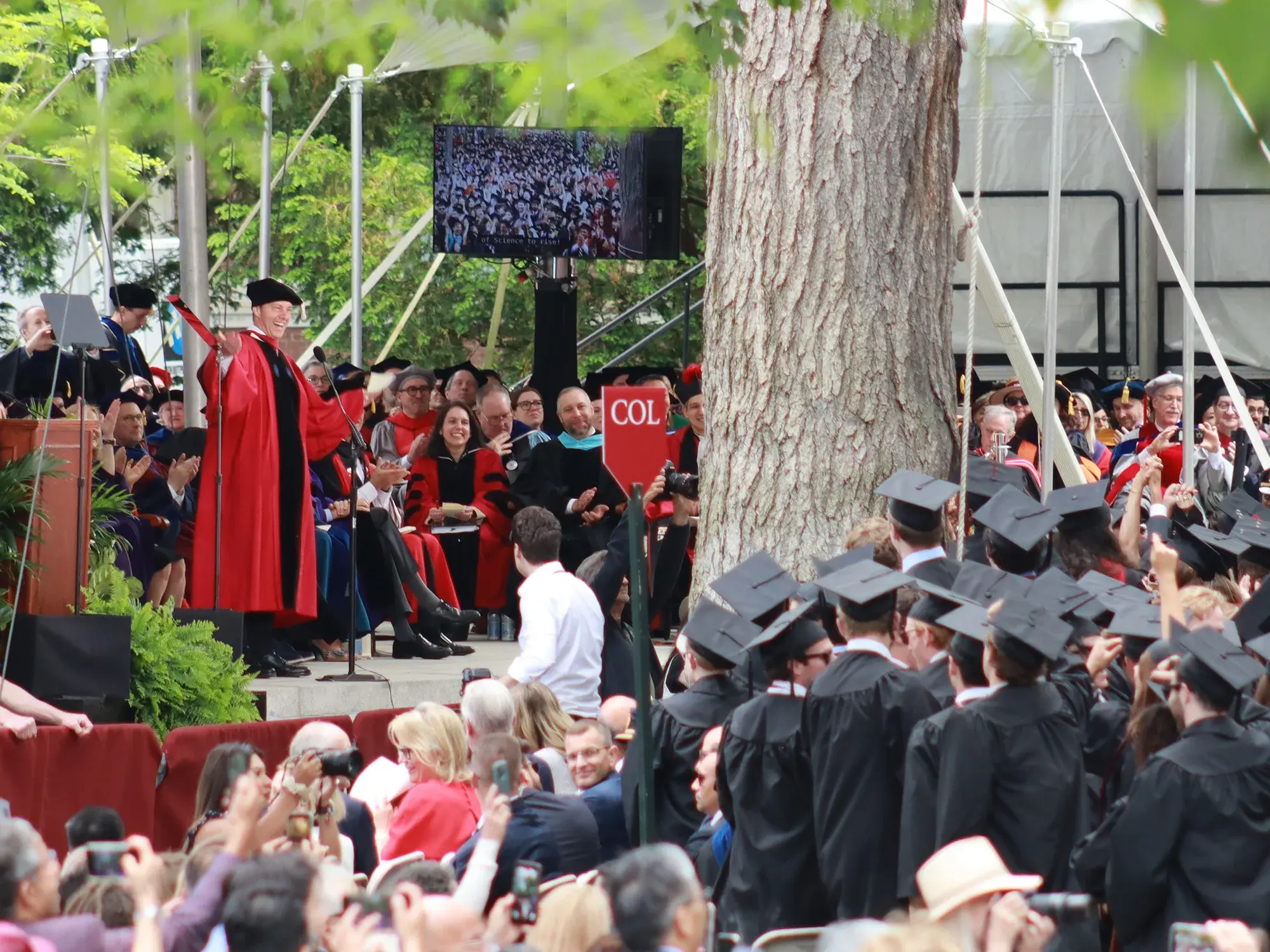 Man in red and black gown and cap speaks to audience of graduates surrounded by trees