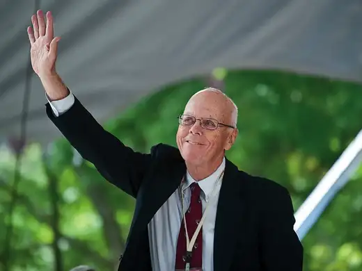 A man in a suit smiles and waves while standing outdoors under a tent.