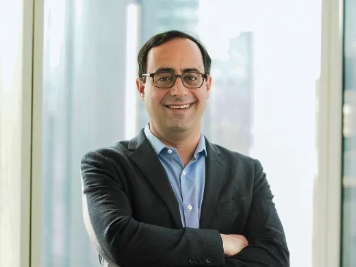 Matt Levine in a dark blazer and glasses stands smiling with arms crossed in front of a large window in a city building.