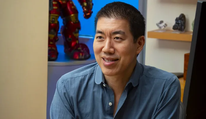 Professor David Liu smiles while sitting at a desk with colorful lanterns and a figurine in the background.