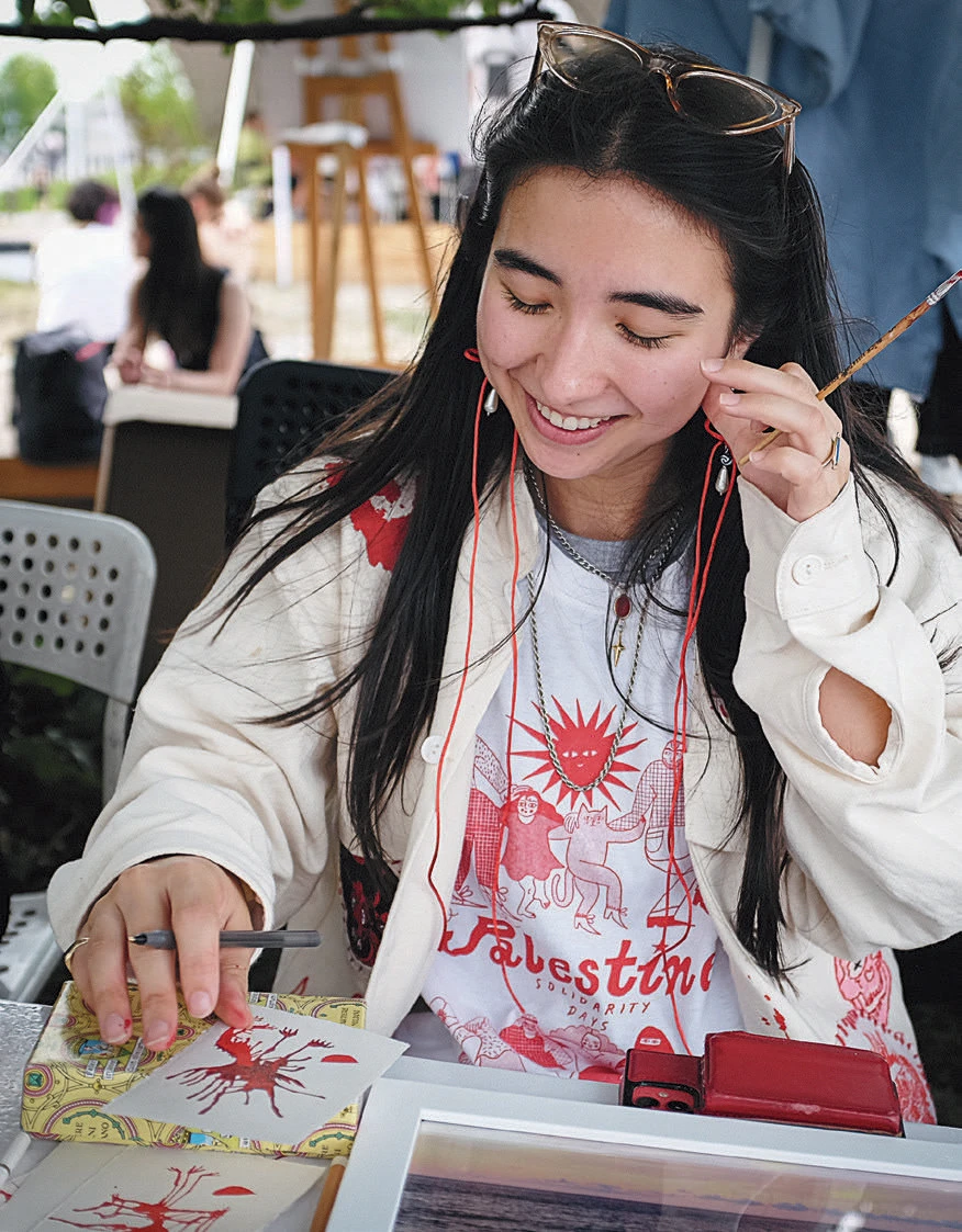 Smiling woman at a table painting a red design while wearing a Palestine solidarity shirt.