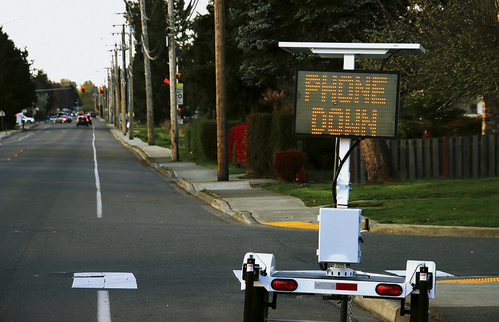 An electronic road sign with the words “PHONE DOWN” stands on a sidewalk near a street lined with trees.