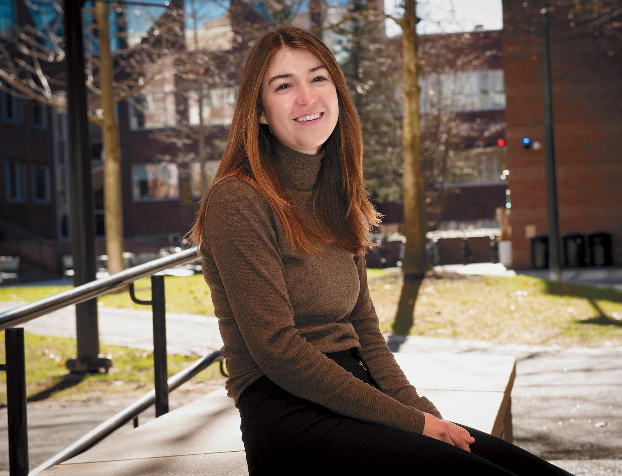 Clare Suter seated outside on a sunny day, smiling with trees and campus buildings in the background