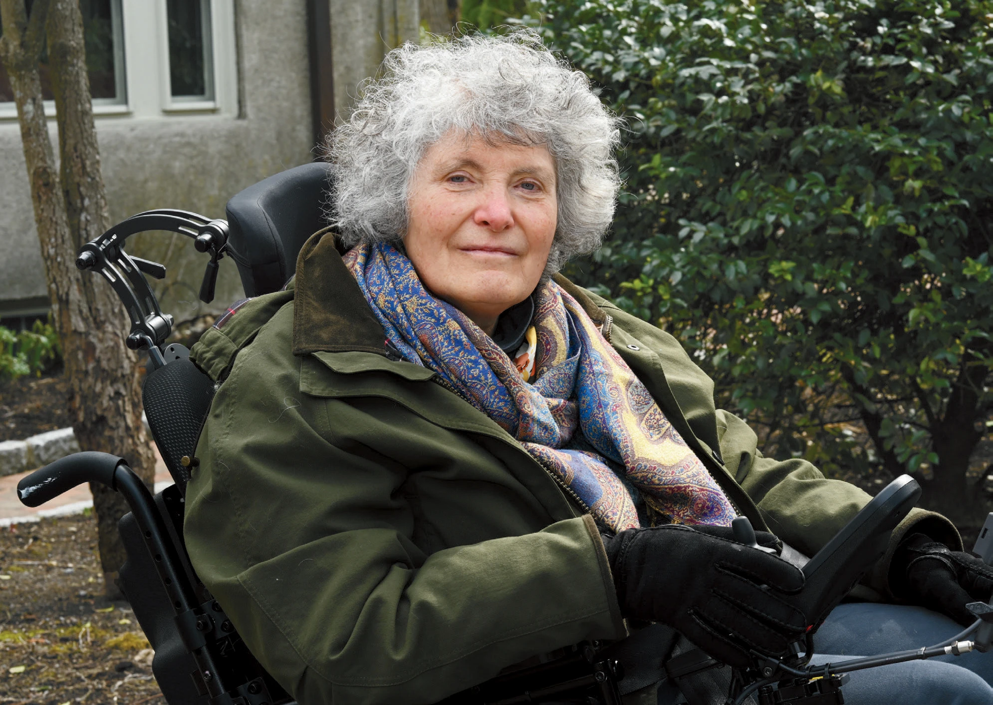 Lisa Iezzoni in a green coat and patterned scarf, seated in a wheelchair beside shrubs and trees