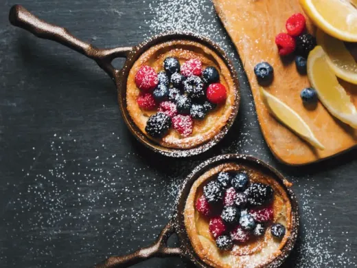Two small cast iron pans with berry-topped desserts, dusted with powdered sugar, alongside lemon slices.