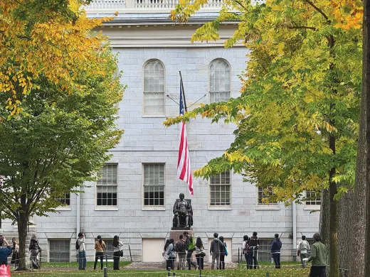People gather near the John Harvard Statue in front of University Hall surrounded by autumn trees.