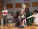 Six women interact in a theatrical setting, one seated and being comforted by others.