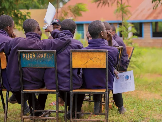 Students in purple jackets seated on chairs, facing away in a grassy area.