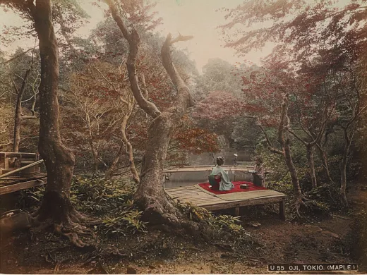 Two women in traditional Japanese clothing sitting on a wooden platform near a tranquil pond, surrounded by autumn foliage.