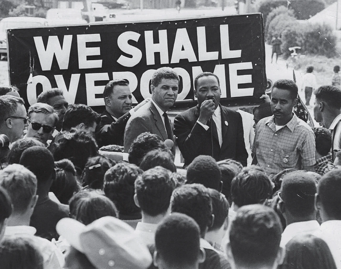 Crowd gathered as civil rights leaders, including Martin Luther King,  speak before a "We Shall Overcome" sign during a protest.